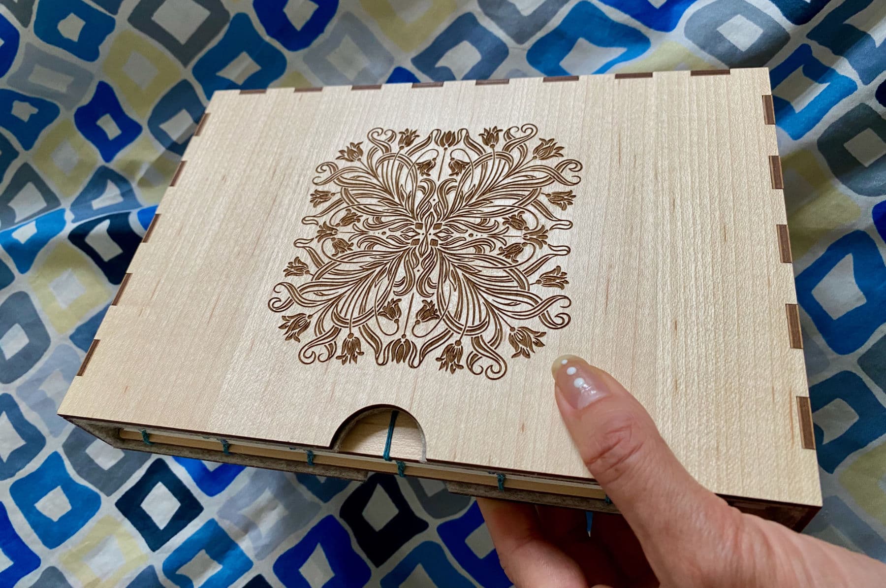 A hand holds a maple wood fondfolio incased in a sturdy slipcase also made from Maple. In the background is a beautiful blue and white patterned vintage scarf which the book will be wrapped in.